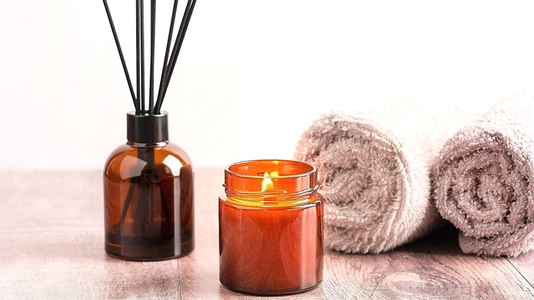 A dark glass candle and diffuser with 2 rolled cream-colored towels on a wooden surface