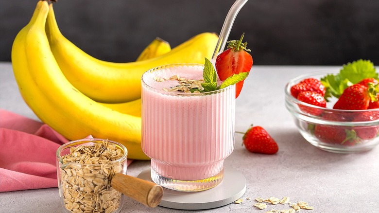 Strawberry smoothie in a short clear glass surrounded by oats, whole bananas, and strawberries