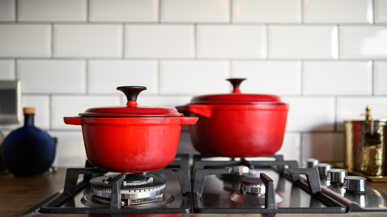 Two vibrant red enamel cast iron dutch oven pots sitting on a modern gas stovetop, set against a backdrop of crisp white subway tiles.