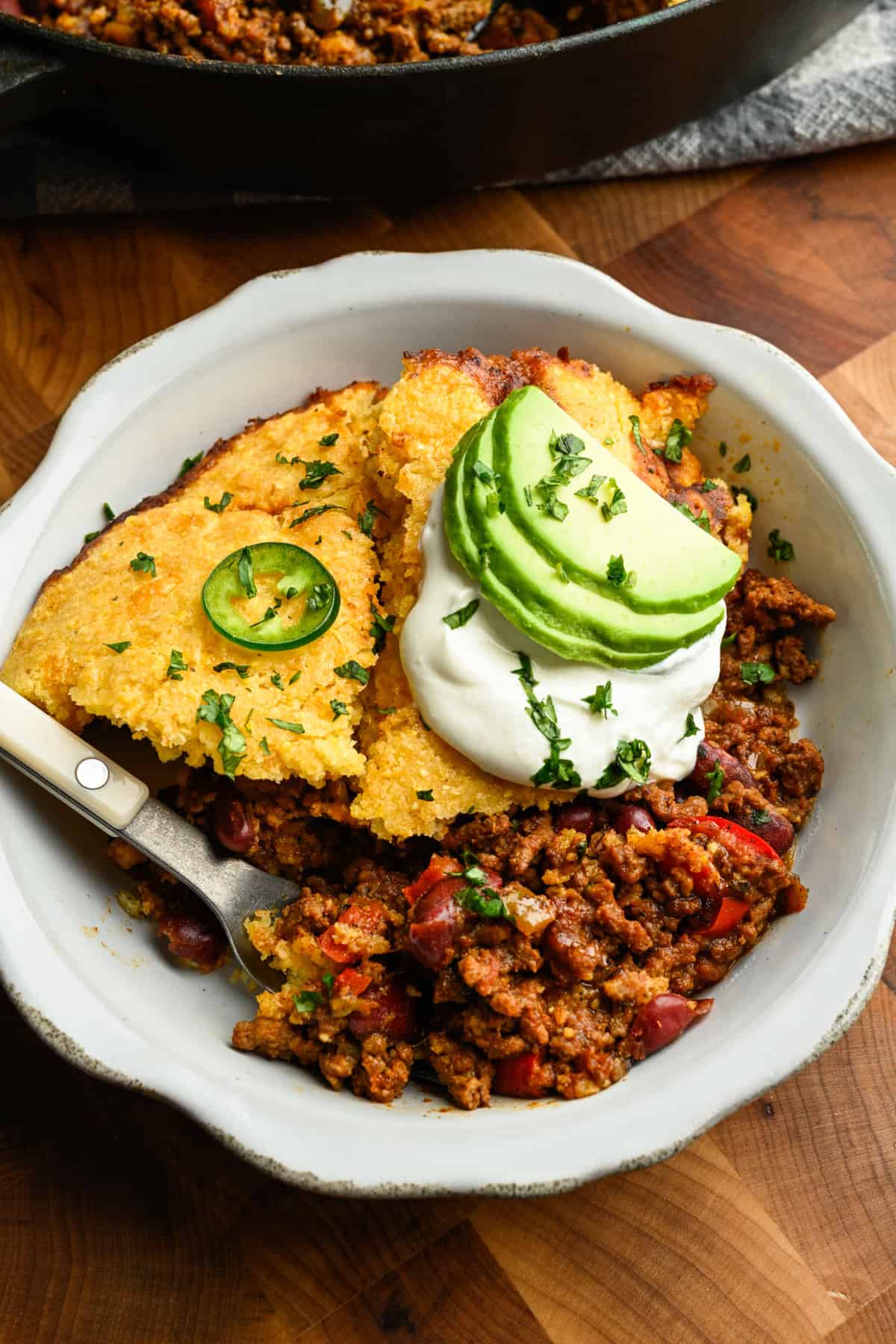 Chili pie topped with cornbread in a white bowl with avocado slices.