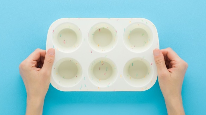 A white silicone muffin baking pan with a colorful sprinkles design held by two hands on top of a blue surface.