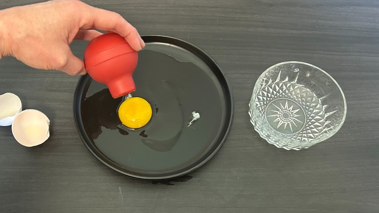 Bulb of a turkey baster sucking up an egg yolk from a black plate next to a glass bowl
