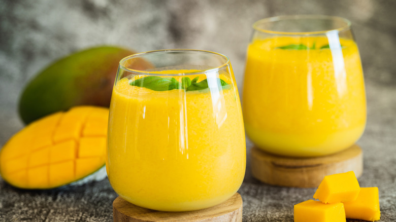 Two glasses of mango lassi with cut fresh mango in the background