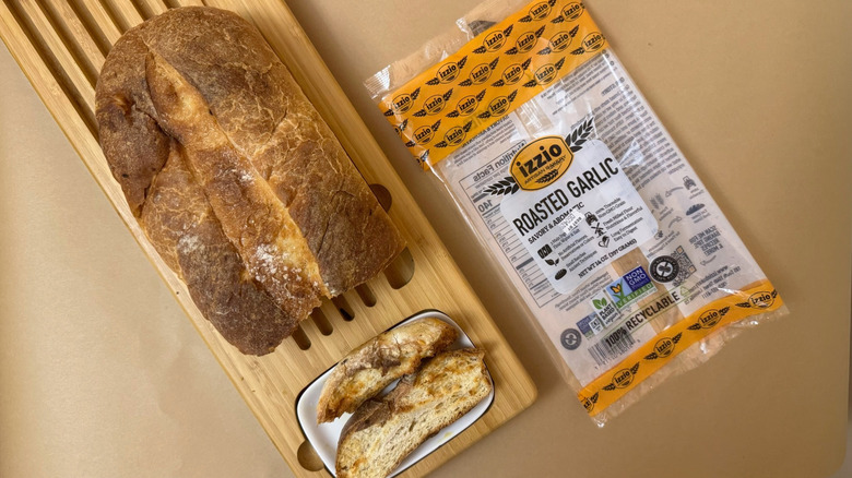 Loaf of Izzio Artisan Bakery Take & Bake Roasted Garlic Sourdough on wooden board beside product packaging