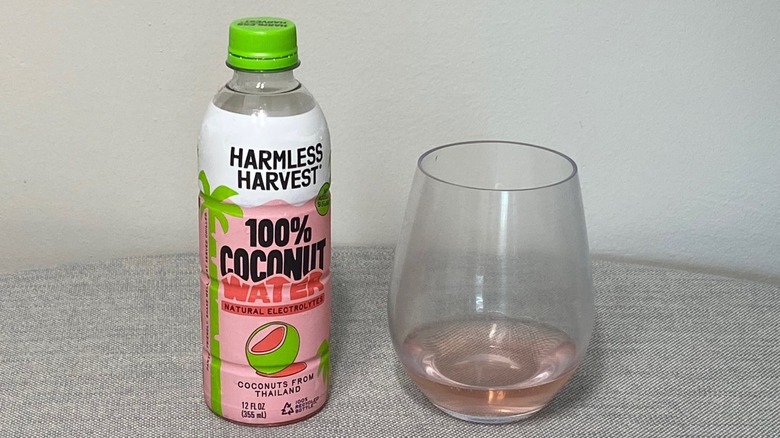 A bottle of Harmless Harvest coconut water on a table beside a glass of the product, which is pink-tinged