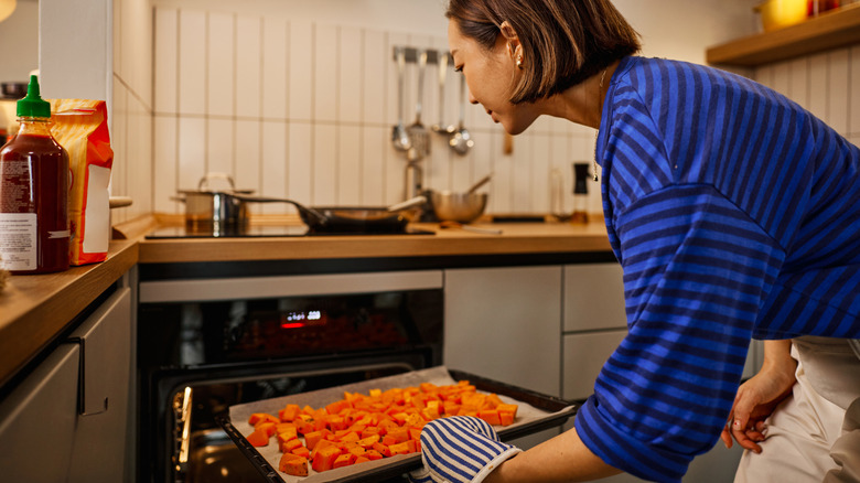 A woman places a sheet pan lined with parchment paper and filled with cubed squash into the oven in a modern kitchen