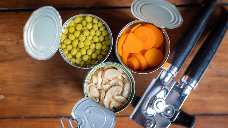 Open cans of peas, carrots, and mushrooms on a wooden table with a can opener
