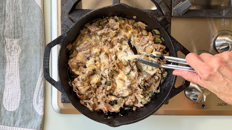 Mixing beef, peppers, and onions with cheese sauce in cast iron skillet