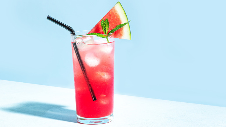A glass of watermelon juice with a bendable black plastic straw, a wedge of watermelon, and a mint leaf garnish