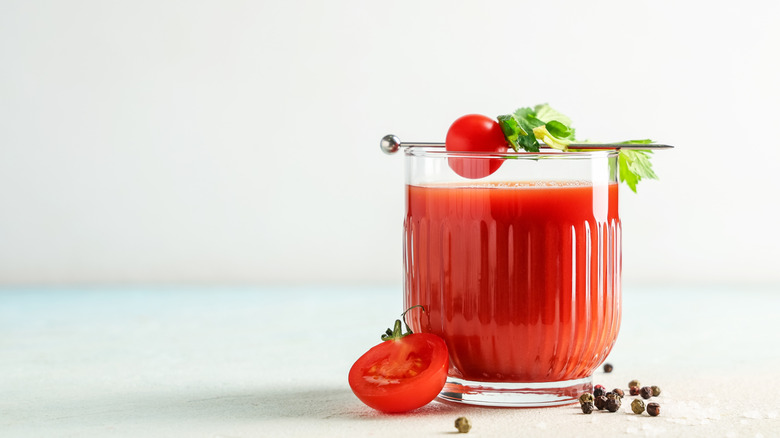 Glass of tomato juice with tomato and celery garnish, next to a halved tomato and some loose peppercorns