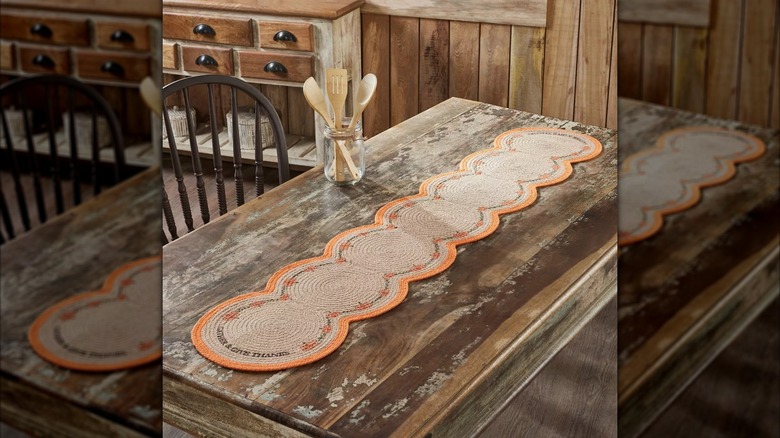 A woven table runner with small pumpkins lining it laying across a wooden table with a jar of kitchen utensils nearby.
