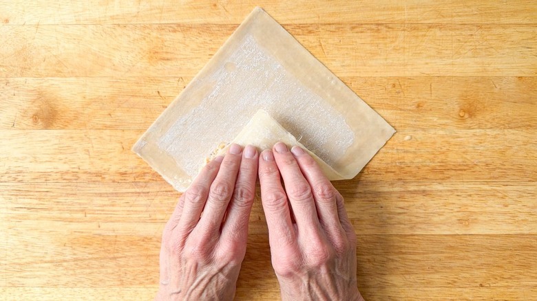 Folding the bottom of an egg roll wrapper over crab cake filling on cutting board