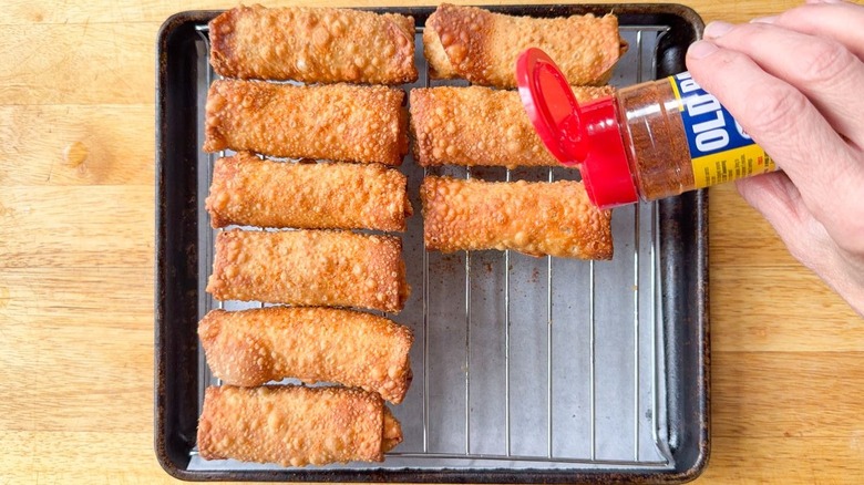 Sprinkling Old Bay seasoning on freshly-fried crab cake egg rolls draining on rack