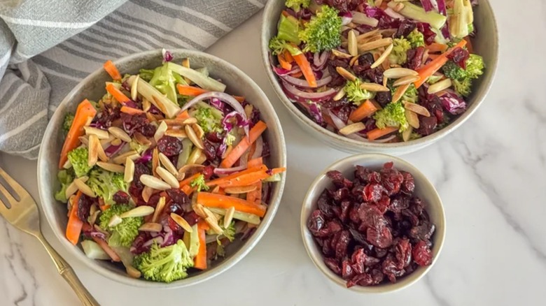 two bowls of sweet and savory broccoli slaw next to a bowl of dried cranberries