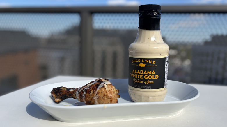 A bottle of Duce's Wild Alabama White Gold next to a chicken leg drizzled with the sauce