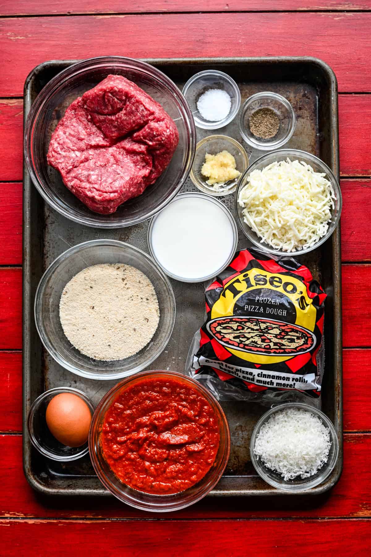 Overhead view af kødbolle-sub bidder ingredienser på en sheet pan: hakket oksekød, brødkrummer, mælk, æg, hvidløg, mozzarella, parmesan, marinara og Tiseos frosne pizzadej.
