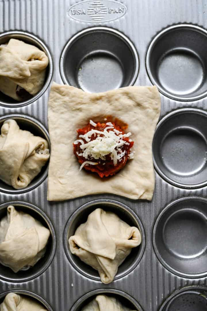 Overhead view af marinara og revet mozzarella skefter på et dejstykke i en muffin-tin.