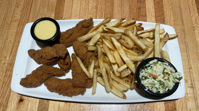 Chicken tenders sit on a plate with honey mustard, fries, and cole slaw.