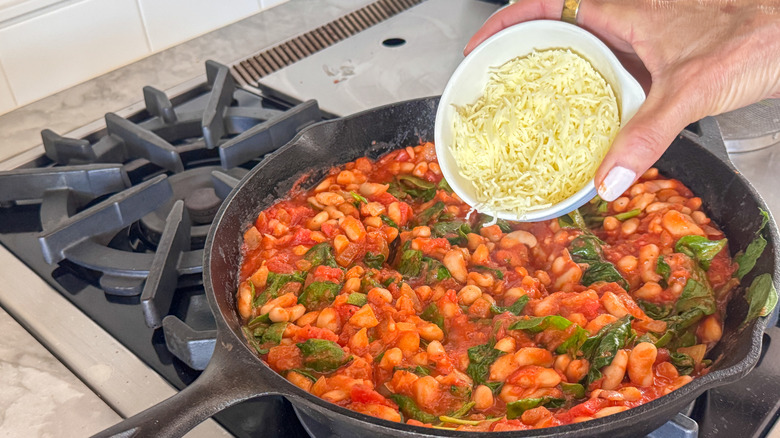 Hand pouring bowl of shredded cheese into pan with beans, sauce, and spinach
