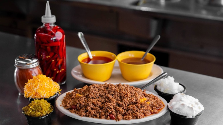A plate of chili with cups of toppings including shredded cheese, relish, onions, sour cream, cups of broth, and chili oil