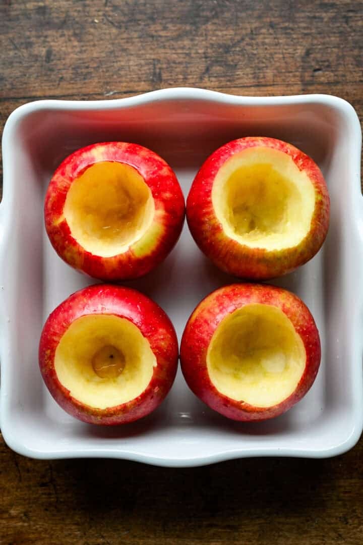 Four honeycrisp apples, cored with the bottoms left intact, standing in a white square baking dish.