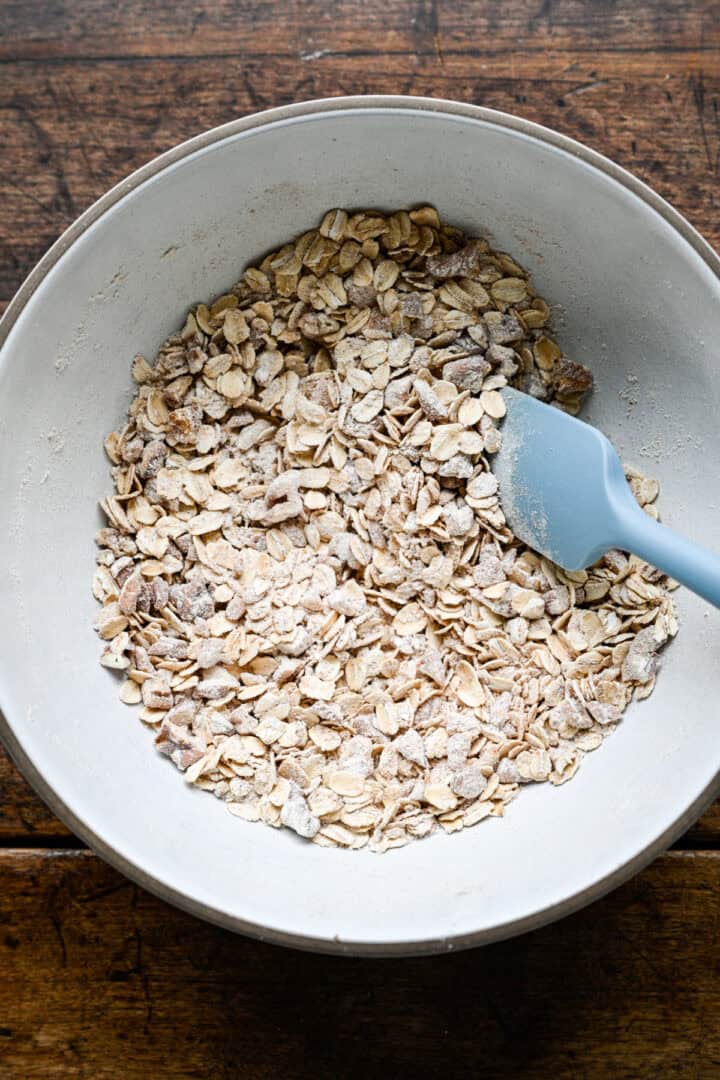 Oats, pecans and crystallized ginger tossed with flour and spices in a white bowl with a blue spatula.
