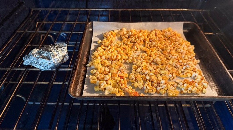 Seasoned corn kernels on parchment-lined baking sheet and garlic wrapped in foil in oven