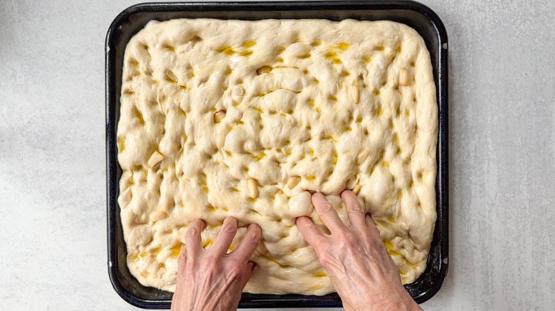 Dimpling focaccia dough topped with olive oil and roasted garlic cloves with fingertips