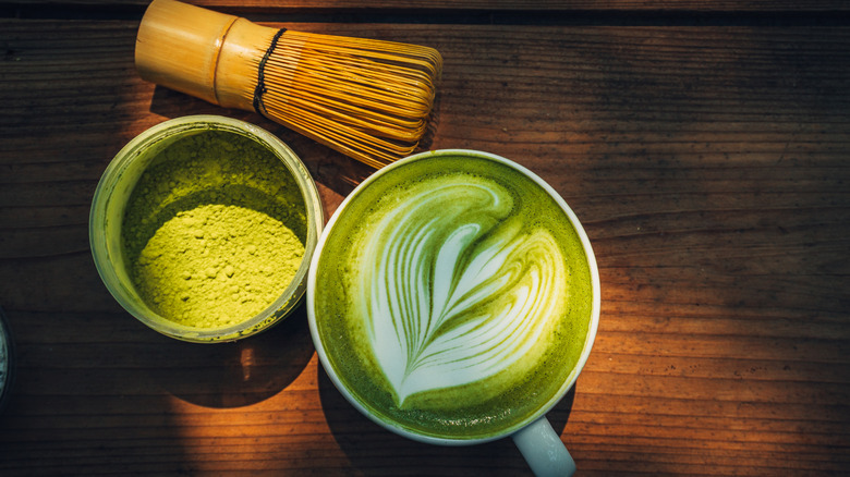 A matcha latte next to a jar of matcha powder and a chasen