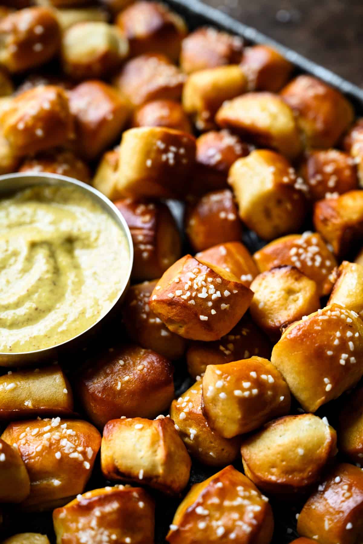 Angled close-up of glossy, salted pretzel bites on a sheet pan next to a bowl of mustard dip, showing the soft, golden sides.
