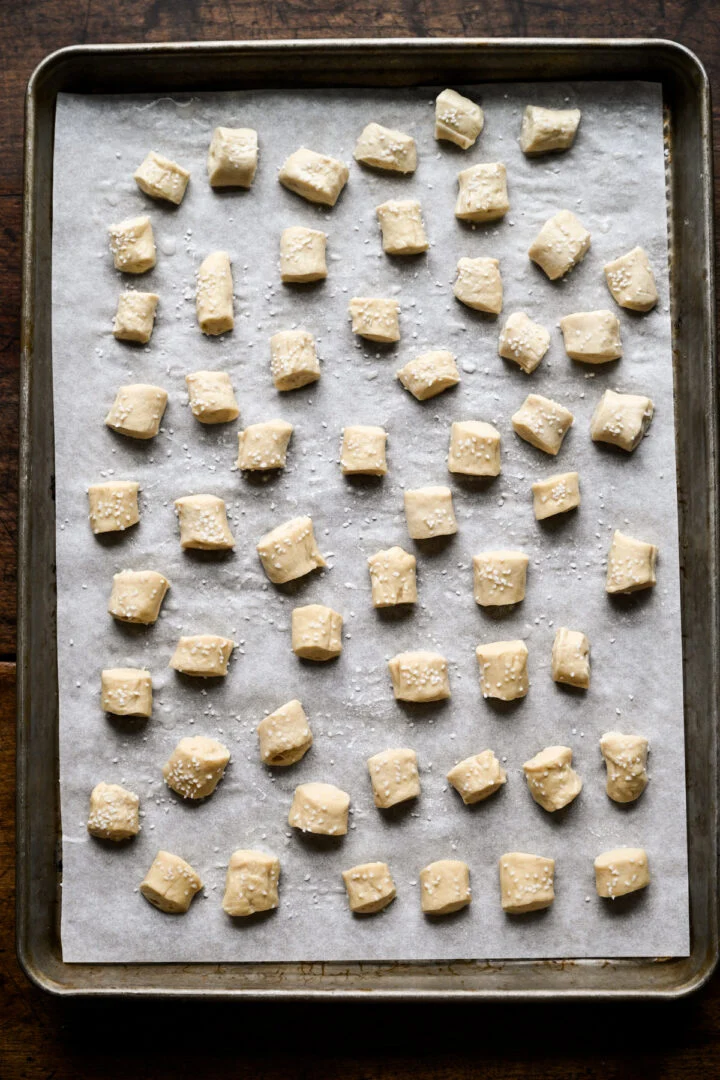 Rows of cut, unbaked pretzel dough pieces sprinkled with pretzel salt on a parchment-lined sheet pan, ready for the oven.