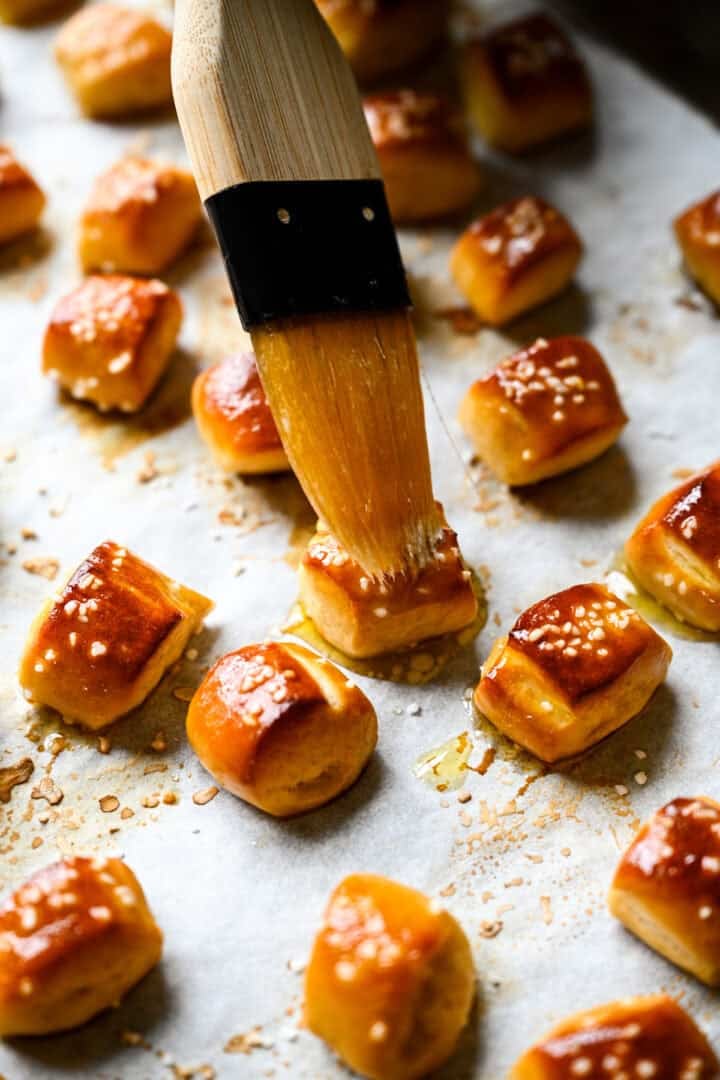 Pastry brush coating freshly baked pretzel bites with melted butter on a parchment-lined sheet pan.