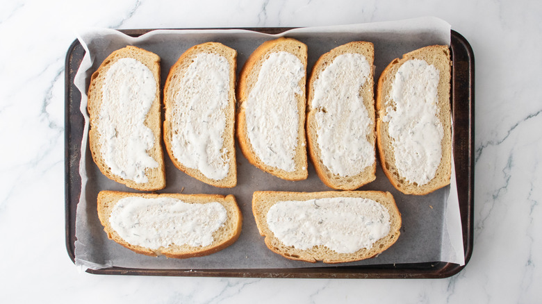 A large baking sheet lined with wax paper containing bread slices spread with ranch dressing.