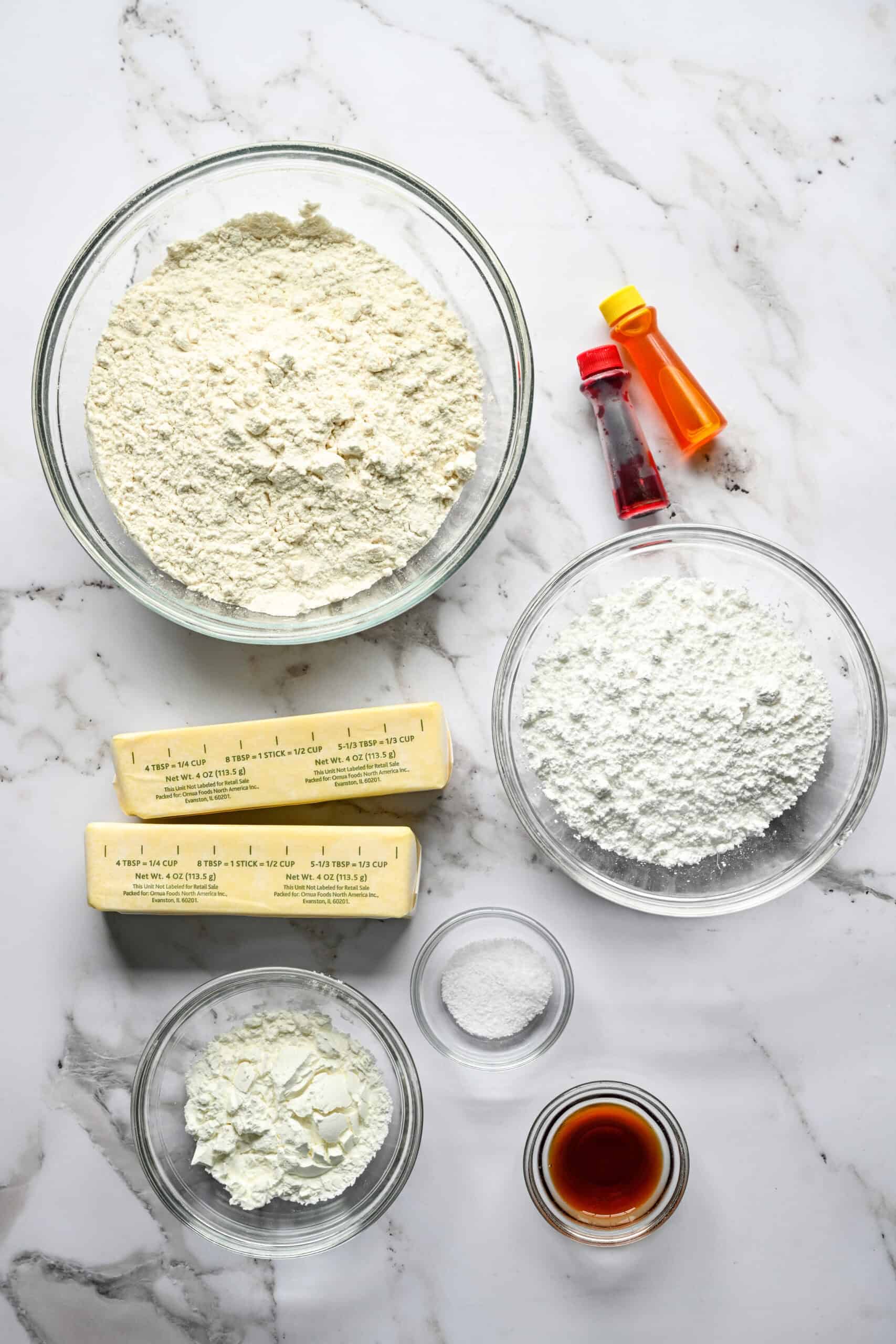 Ingredients for candy corn shortbread cookies on a marble counter: flour, powdered sugar, two sticks of butter, cornstarch, salt, vanilla and two bottles of food coloring.