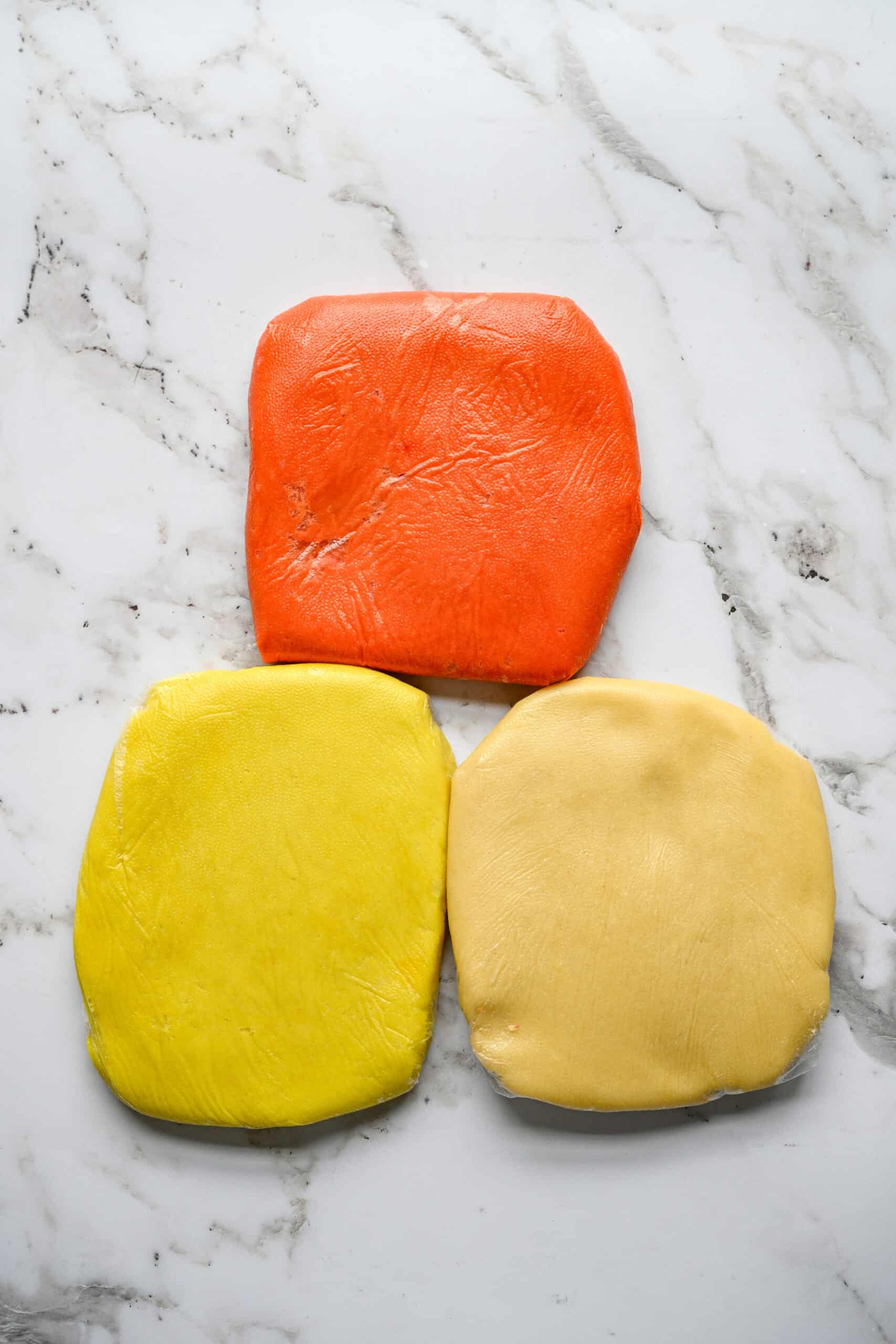 Three flattened portions of shortbread dough on a marble counter: one orange, one yellow and one left plain.