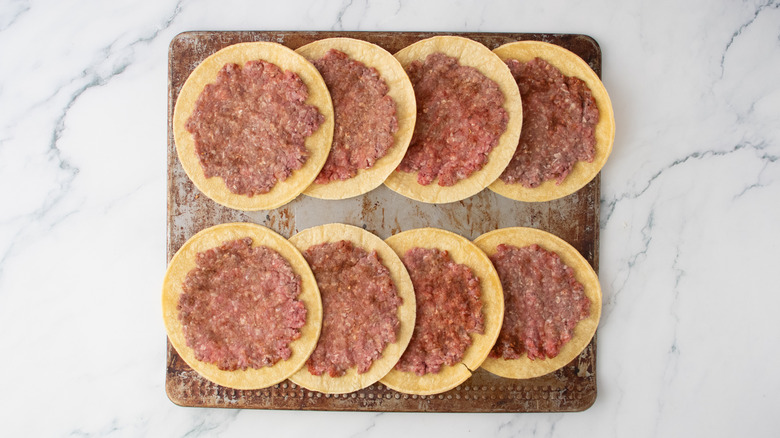 A cookie sheet filled with corn tortillas with smashed burger patties on top