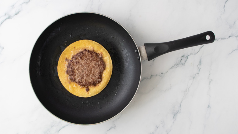 A seared burger patty on a corn tortilla in a nonstick skillet