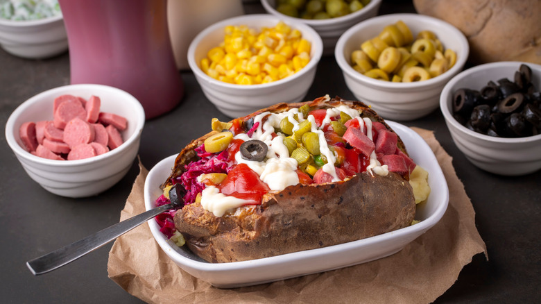A loaded Turkish baked potato surrounded by small bowls of olives, corn, and sausage.
