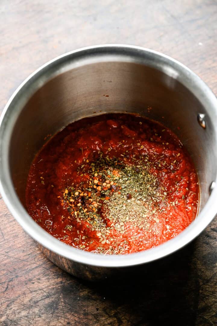 Pizza sauce in a small saucepan topped with Italian seasoning, garlic powder and red pepper flakes.