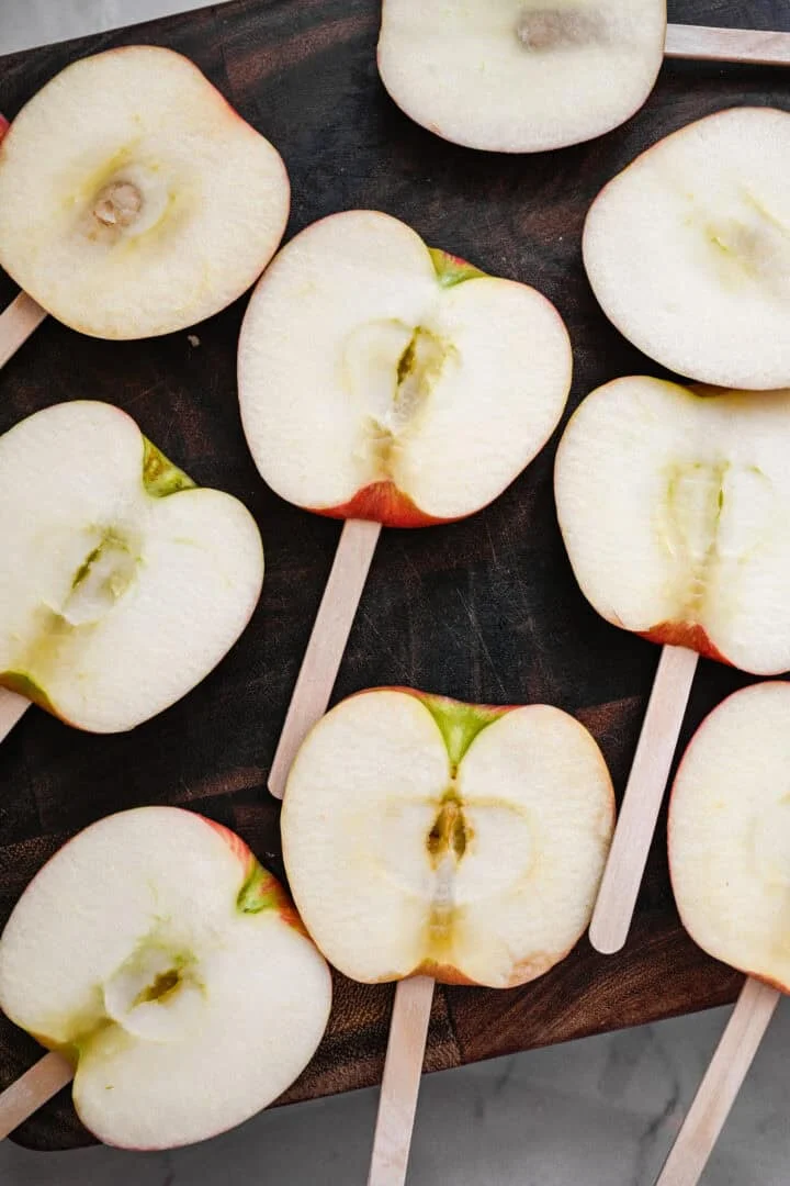Overhead view of thick apple slices on popsicle sticks on a dark wood cutting board.