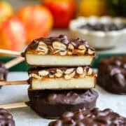 Front view of two halved chocolate covered apple slices stacked to show layers of apple, date spread and peanuts, with apples behind.