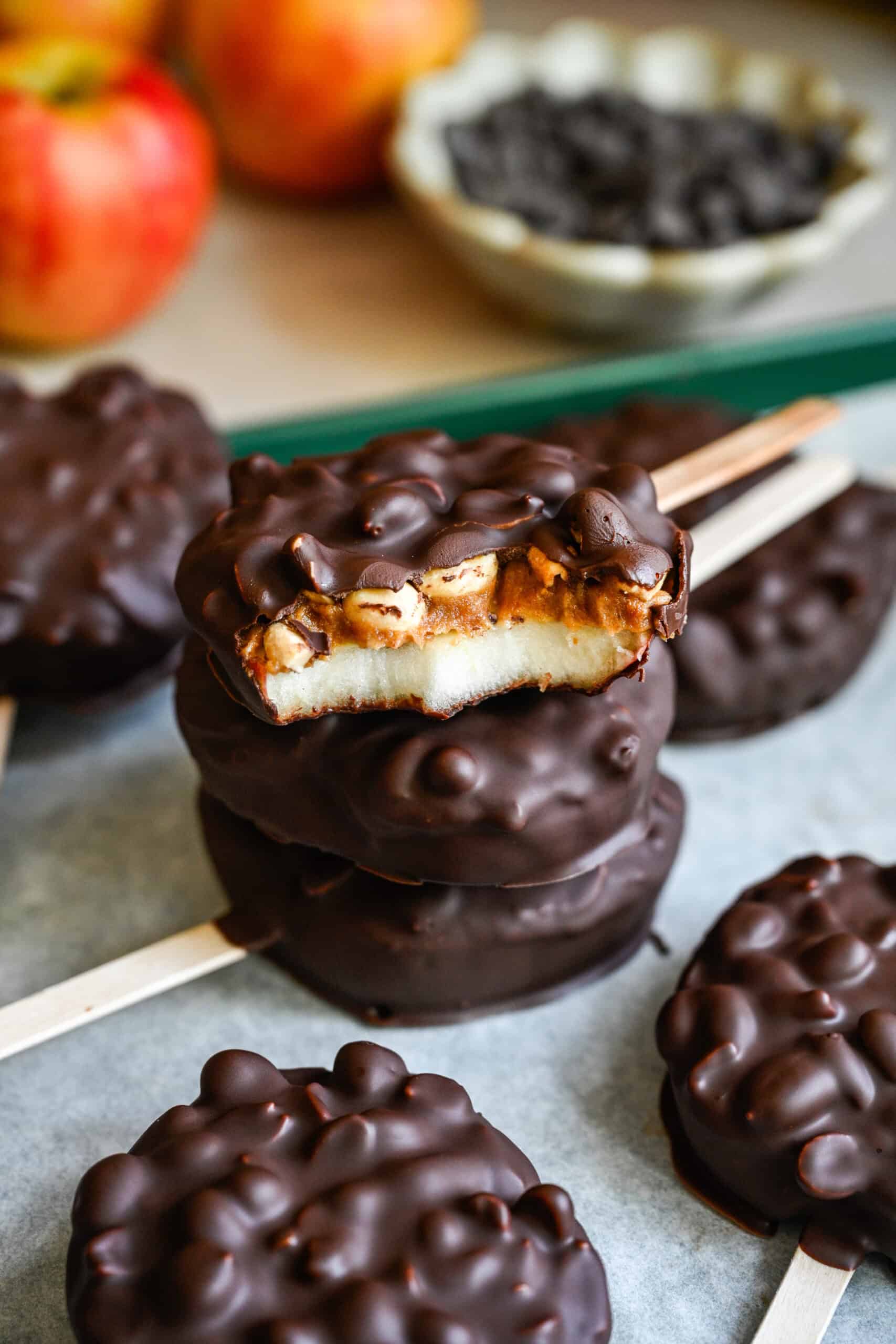 45 degree view of a stack of chocolate covered apple slices, the top one bitten to show the apple, date spread and peanuts.