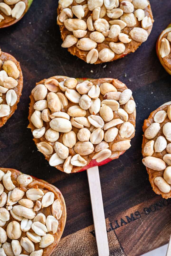 Close up view of apple slices on popsicle sticks topped with date spread and a layer of roasted peanuts.
