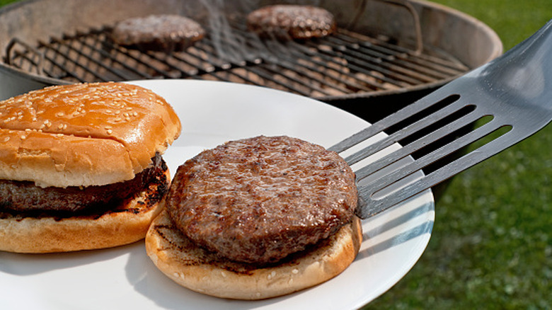 Spatula placing a hamburger onto a bun on a white plate with a charcoal grill in the background