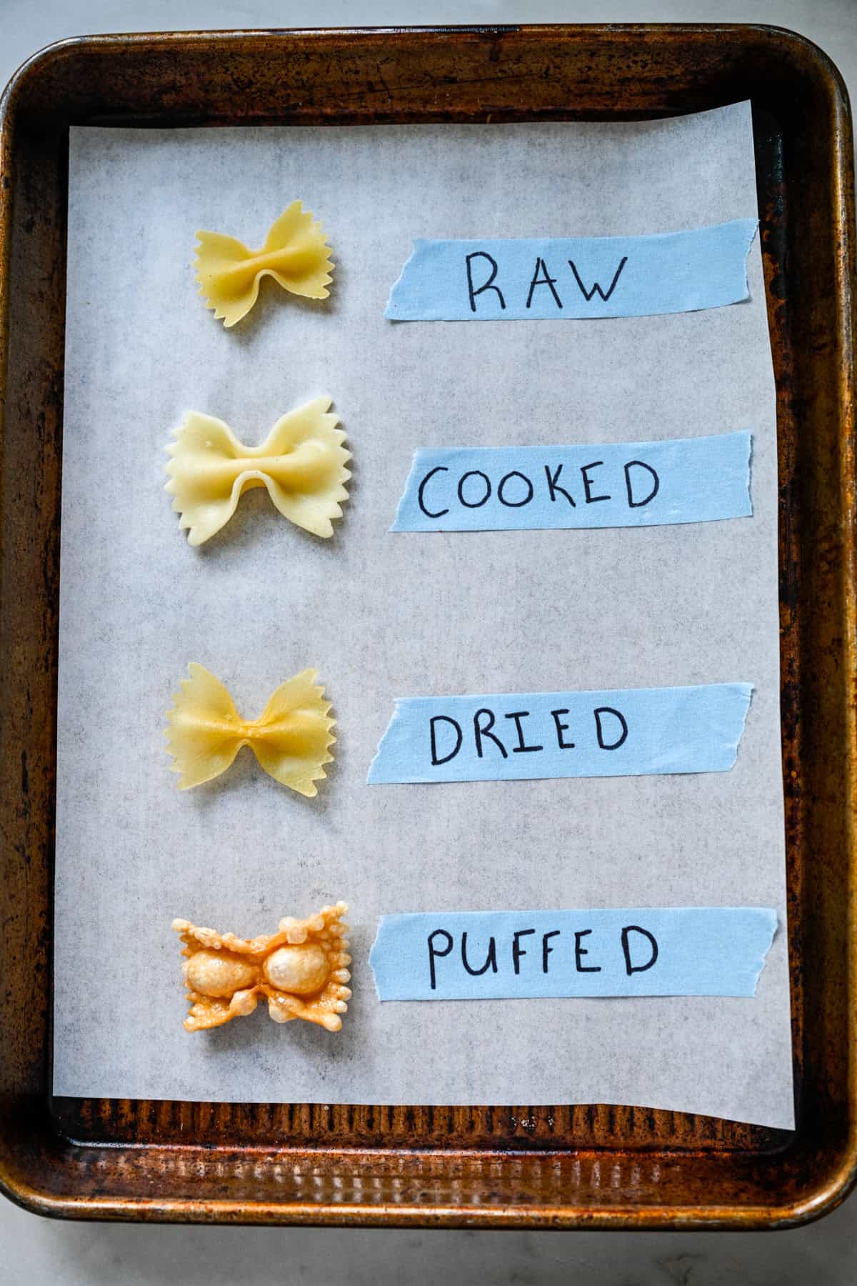 Overhead view of four bow ties on a sheet pan labeled raw, cooked, dried and puffed.