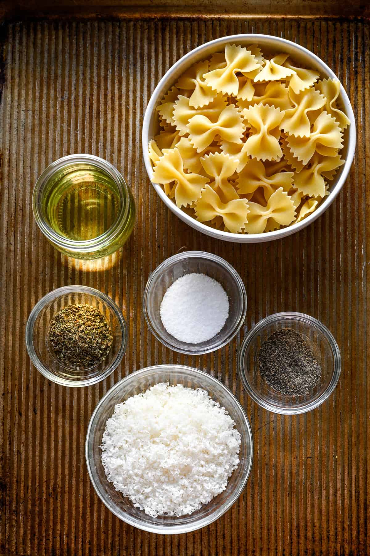 Overhead view of pasta puff ingredients on a sheet pan: bow tie pasta, oil, dried herbs, salt, pepper and grated parmesan.