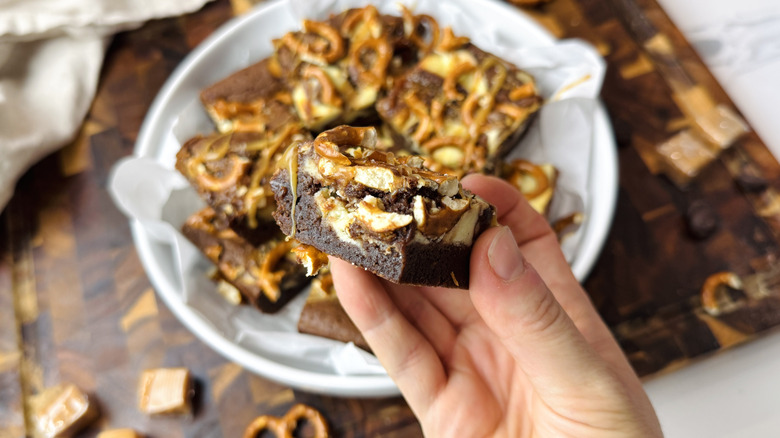 Hand holding caramel pretzel brownie in front of plate with more brownies