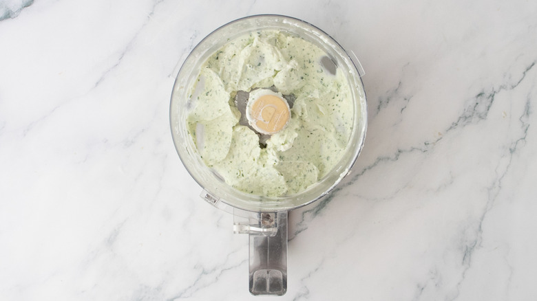 A food processor bowl filled with feta, yogurt, olive oil, and fresh herbs