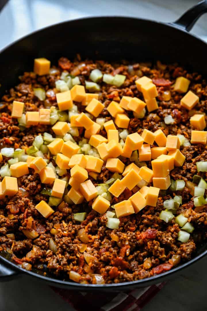 Close up view of cubed cheddar and chopped pickles added on top of the ground beef and bacon filling in a cast iron skillet