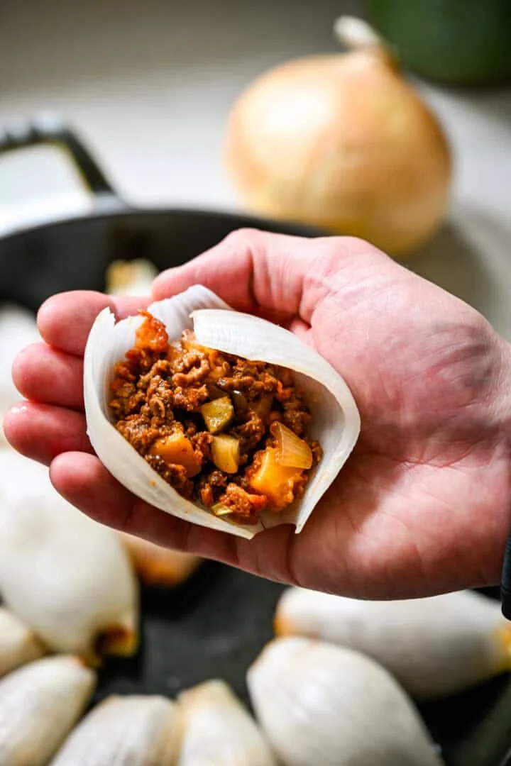 Close up view of a hand holding a boiled onion petal filled with cheeseburger filling before rolling it closed